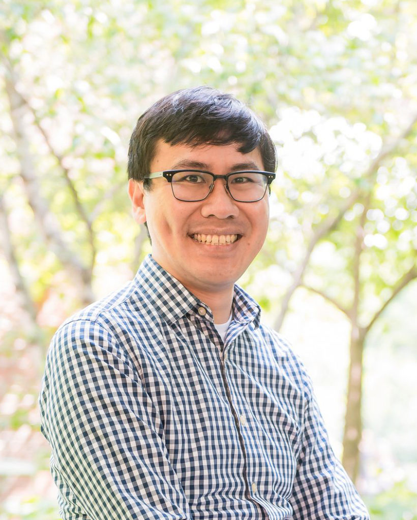 Young east asian man smiling in front of a background of trees. He is wearing black rimmed glasses and a blue gingham button up shirt.